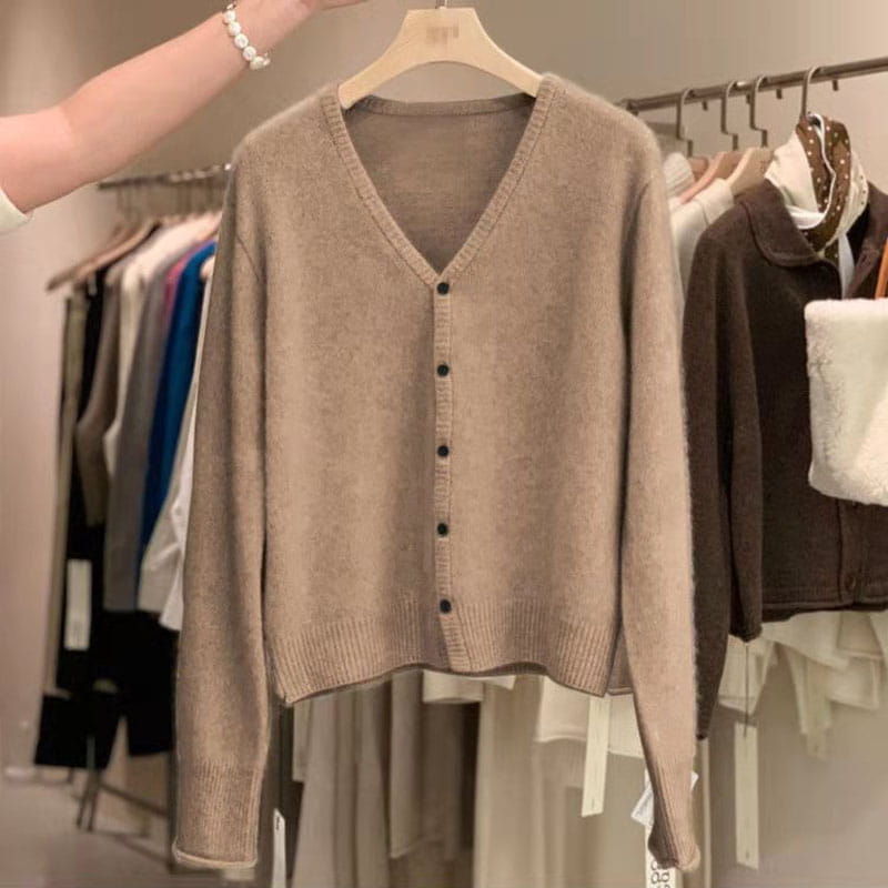 Un pull cardigan en laine mérinos beige doux à encolure en V, avec des boutons noirs, suspendu à un cintre en bois.