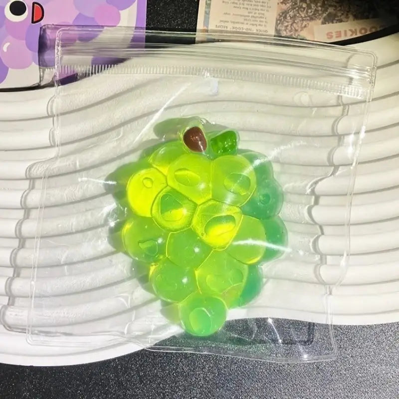 Ein transparenter, grüner und gelber Trauben-geformter Gel-Quetschspaß mit einem kleinen braunen Stiel, eingehüllt in eine transparente Kunststoffverpackung für taba-licious Fun Fruit Fidget-Spiel.