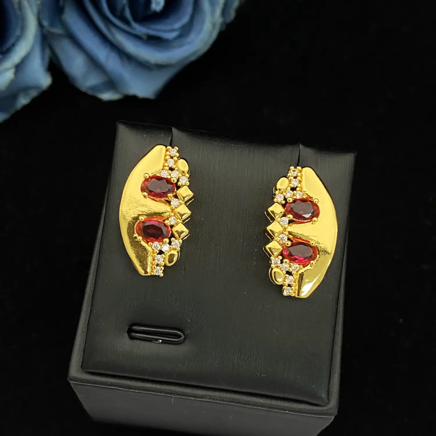 Ensemble de bijoux géométriques en zircon doré comportant des boucles d’oreilles de couleur dorée en forme de feuille, ornées de pierres ovales rouges et de cristaux clairs entourants.