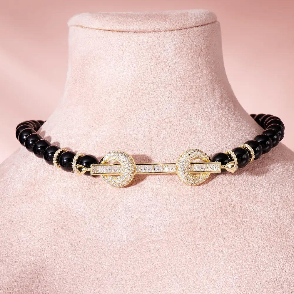 Elegante collar tipo choker con cuentas negras, con un broche de tono dorado engastado con diamantes, elaborado con joyería de circonita cúbica.