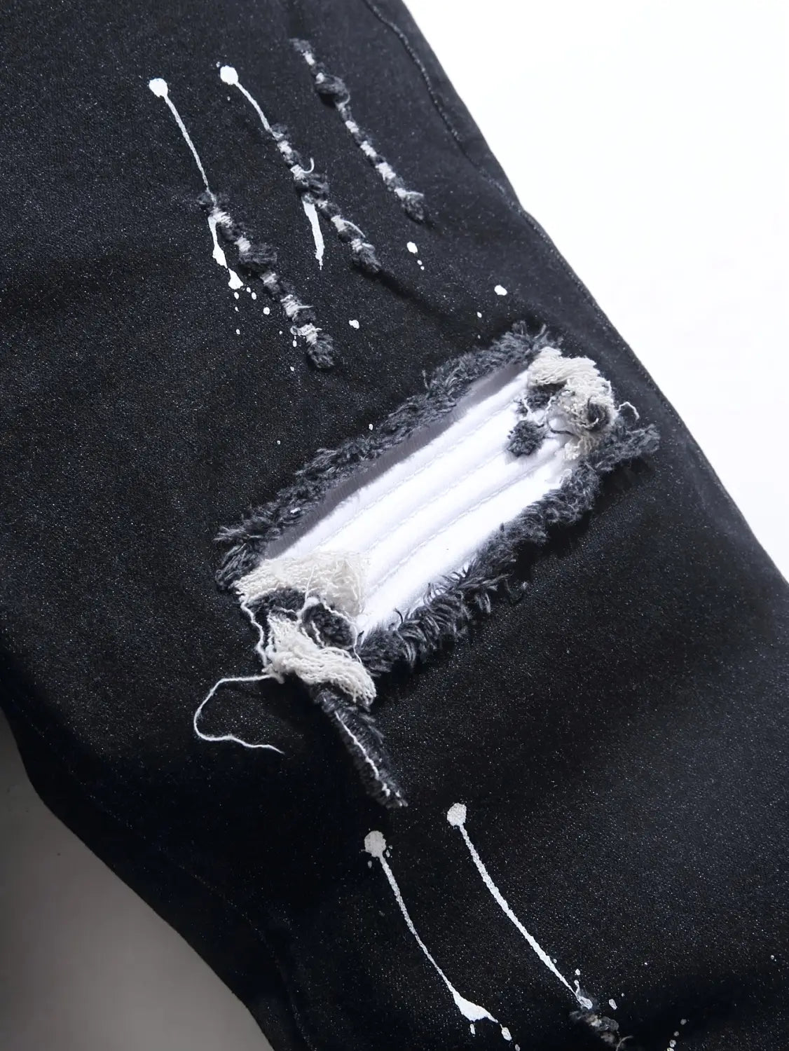 Herren-Ripped-Skinny-Bikerjeans grau mit besticktem Streetwear-Design, aus schwarzem abgetragenem Denim mit einer ausgeprägten rechteckigen Aufreißung, ausgefransten Kanten und weißen Farbspritzer-Akzenten.