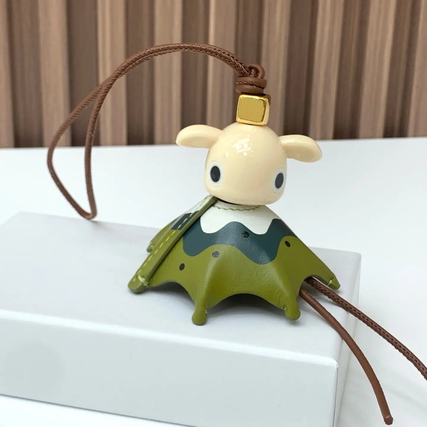 Un llavero colgante para bolso con encanto de animales de cerámica color crema con una base en forma de montaña verde y detalles de puntos negros, diseñado como un llavero tierno.