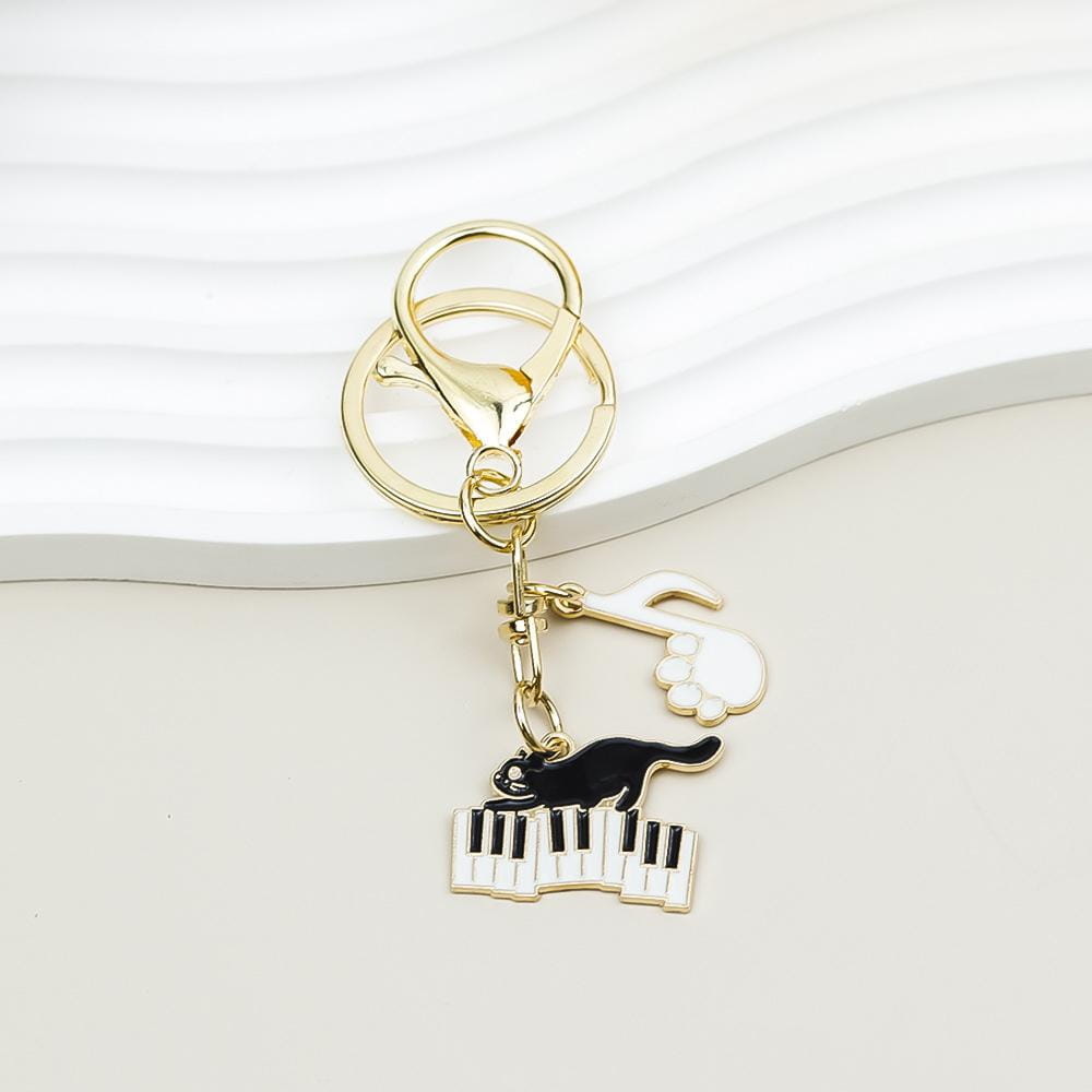 Gold-toned black cat piano keychain featuring a cat perched on piano keys with a white paw print charm above, ideal as a music lover gift.