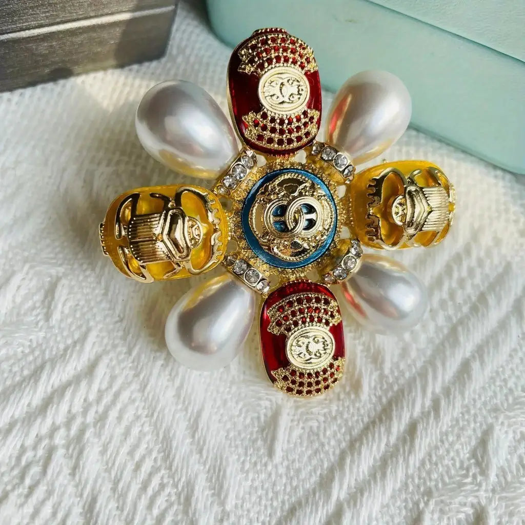 Épingle sertie de rhinestones en forme de coléoptère pour broche de perles designer, présentant un médaillon en émail bleu Chanel de qualité supérieure, avec des éléments entrelacés à ton doré, entouré de motifs de coléoptères en émail rouge et jaune, accentués par des rhinestones et posés sur un fond de perles blanches.