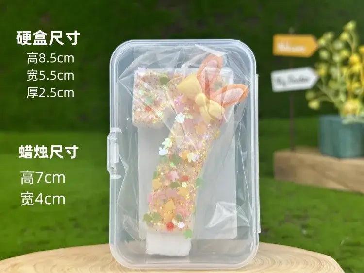 A clear plastic container holds a glitter bunny ear number birthday candle with pastel pink and yellow bunny ears.