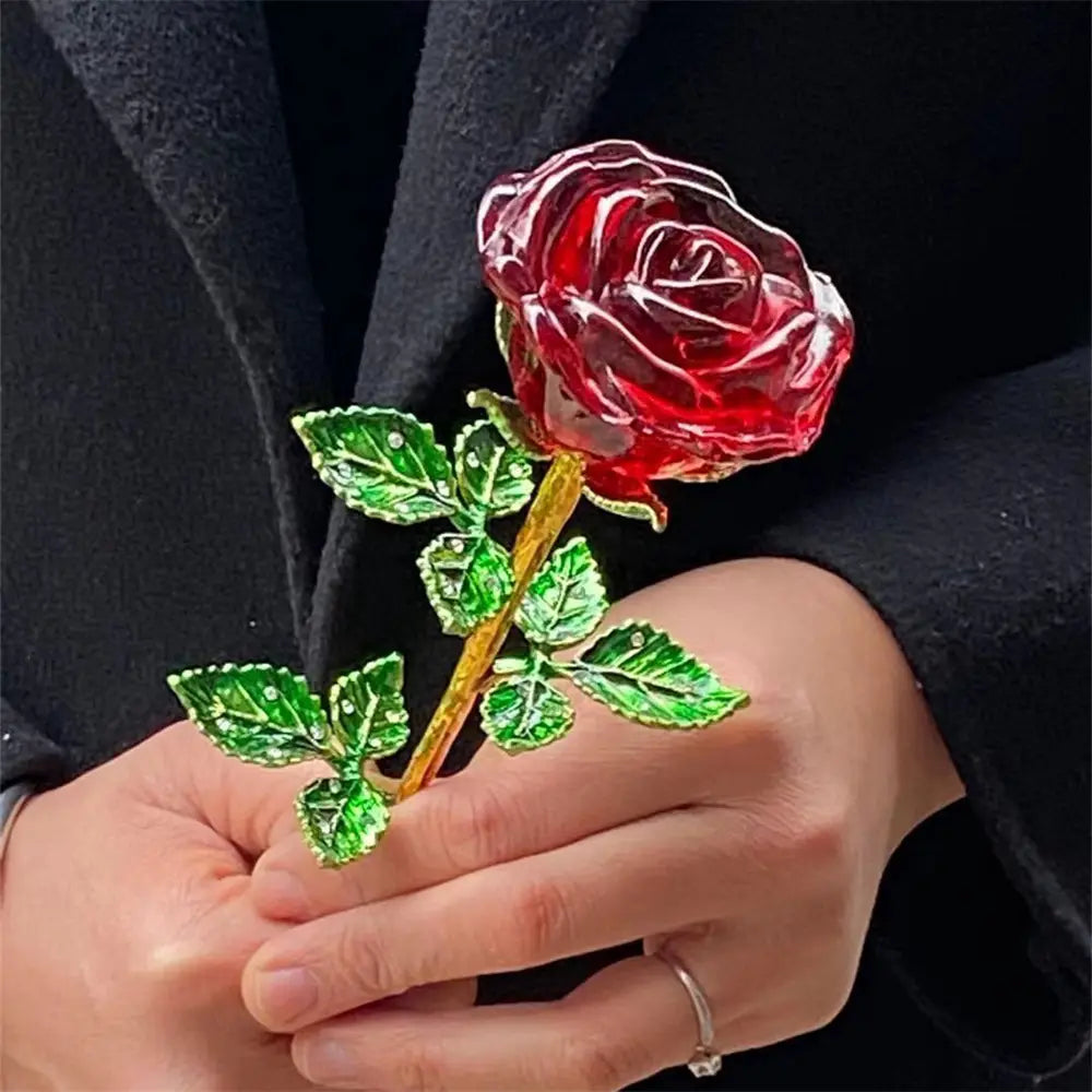 A translucent red crystal glass rose with green enamel leaves and a gold toned stem held delicately in hands ideal as a Valentine’s Day gift.