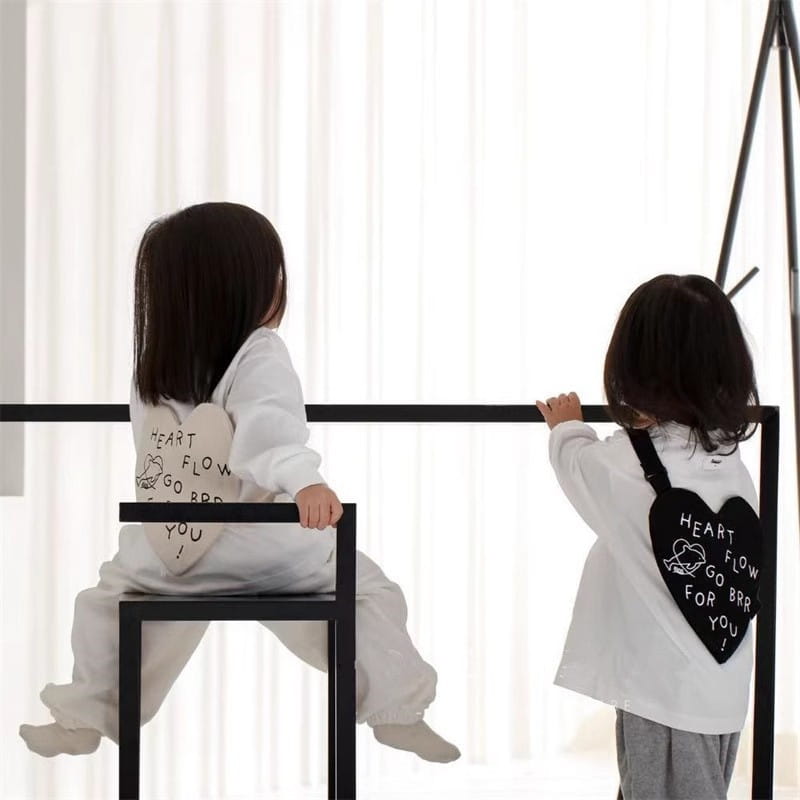 Weiße herzförmige Kinder-Umhängetasche aus Korea mit schwarzer Beschriftung und kleinem Cartoon-Design auf der Vorderseite.