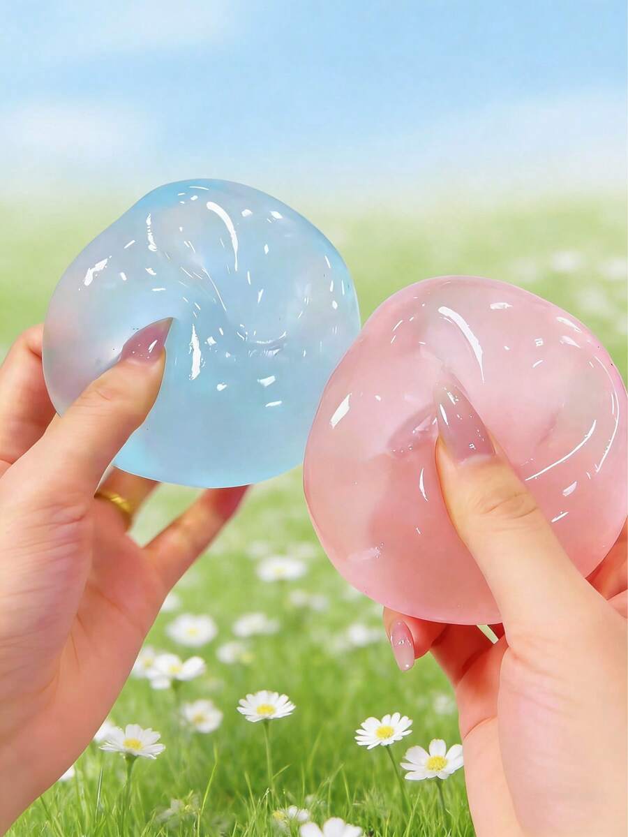 Deux balles anti-stress translucides et souples, bleu clair et rose tendre, tenues délicatement dans les mains, contre un arrière-plan flou de marguerites blanches, pour le produit « Blue Coconut Oil Stress Ball Squeeze Toy ».