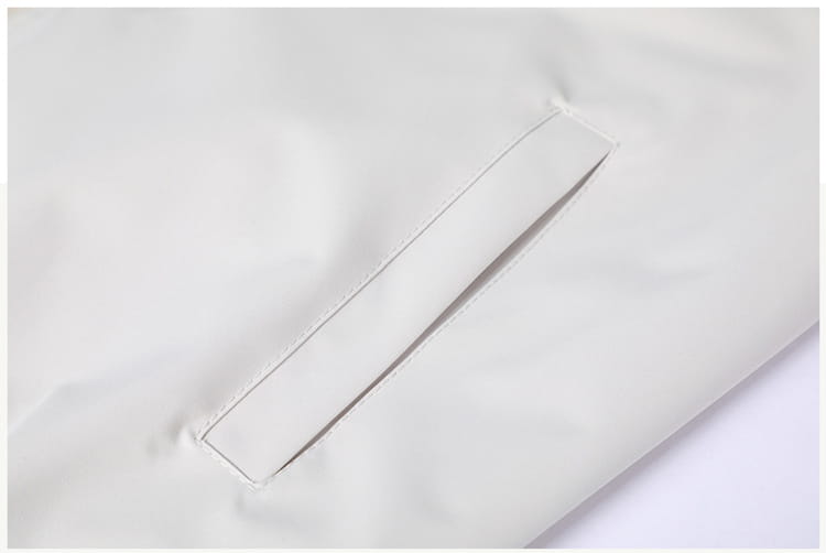 Un bolsillo de tejido color crema pálido con costuras limpias y un brillo sutil en una chaqueta cortavientos ligera para hombre con cuello vuelto.