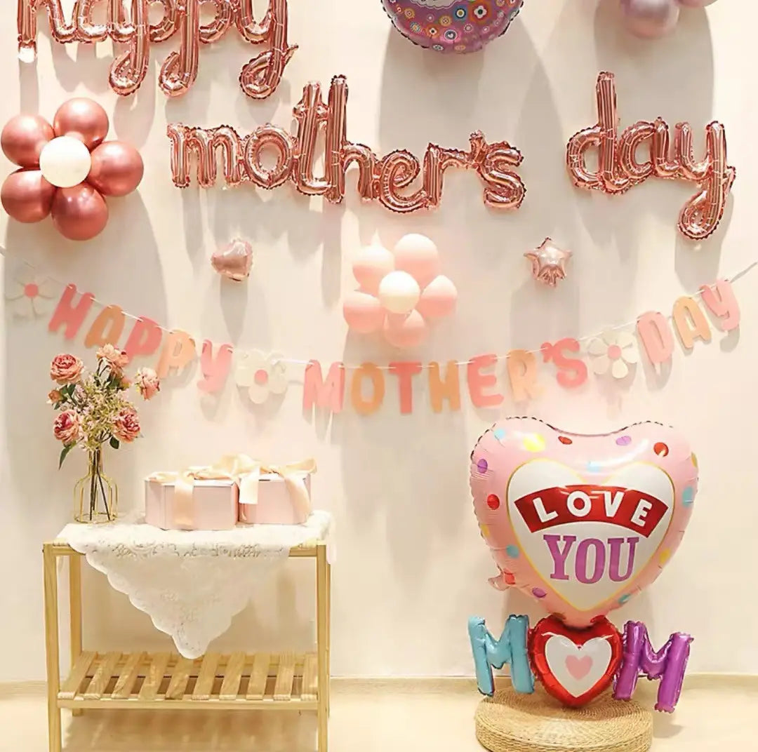 a mothers day daisy paper banner wall decor featuring a pink heart-shaped balloon with colorful polka dots and a red love you banner standing on a woven base beside purple letter balloons spelling mom