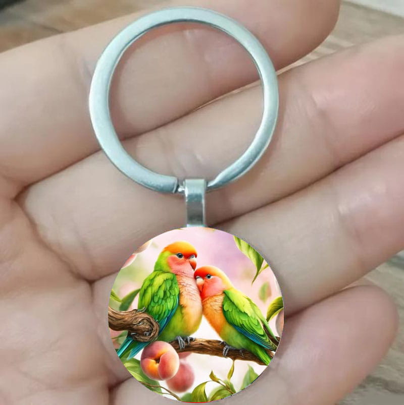 A circular lovebird keychain featuring a vibrant green and orange illustration of two birds perched on a peach branch set in a silver-toned glass dome metal ring.