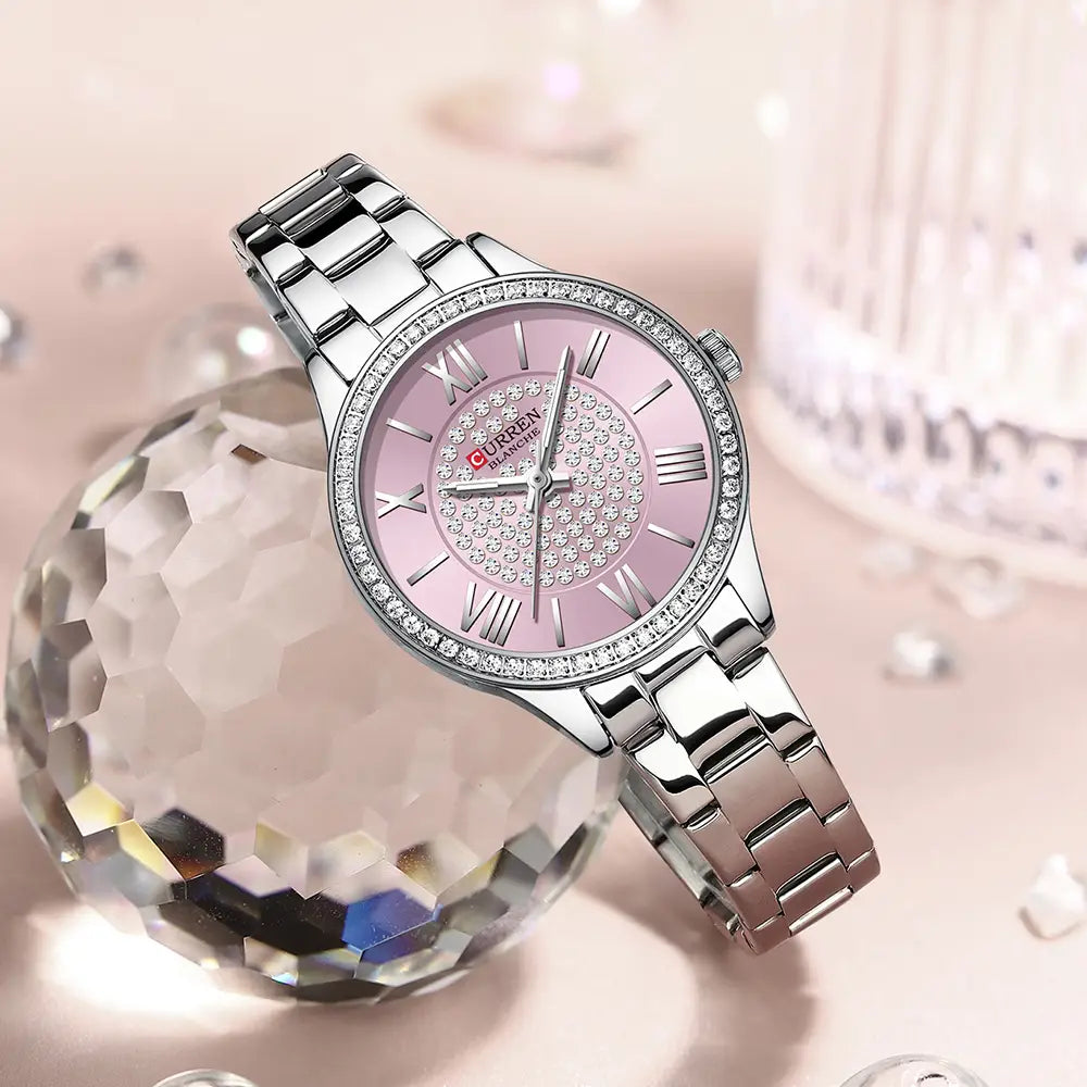 A sleek silver-toned Curren women stainless steel crystal quartz watch with a pink dial featuring sparkling crystals and roman numerals and an elegant metal bracelet.