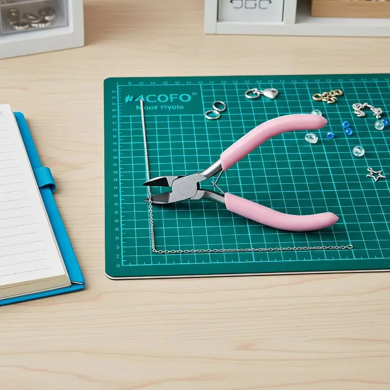 Pinces à bijoux à poignée rose en métal reposant sur un tapis de découpe vert à côté d’une chaîne et d’accessoires de perles assorties pour le travail et les réparations