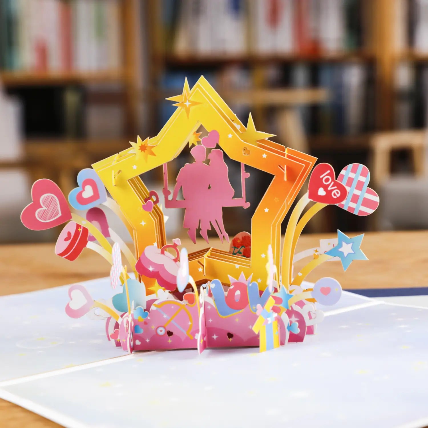 Carte de la Saint-Valentin 3D à effet pop-up vibrante, présentant une silhouette rose d’un couple sur une balançoire, à l’intérieur d’un cadre jaune en forme d’étoile, ornée de cœurs et d’étoiles colorés.