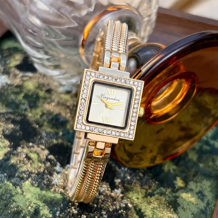 Gold diamond square watch with a gold-toned case, diamond-studded bezel, roman numeral markers, and cream-colored dial on a vintage fringe strap.