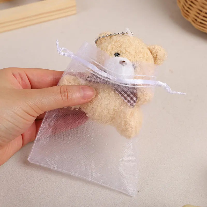 白い顔、黒いボタンの目、茶色のチェック柄の蝶ネクタイを備えた柔らかなベージュのぬいぐるみテディベアが、同じリボンで結ばれたシアーなオーガンジーのギフトバッグの中に保持されており、オーガンジー袋付きのテディベアのお祝品セットの一部として用いられます。