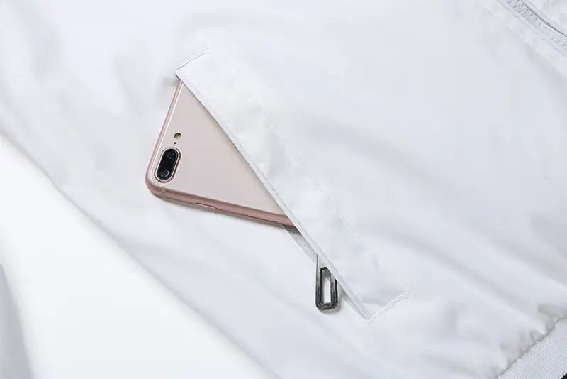 Un iPhone 7 Plus color rosa dorado colocado dentro del bolsillo de tela blanca, con tirador de cremallera plateado de la chaqueta tipo bomber USA para hombre, con gráfico de letras estilo varsity, abrigo casual