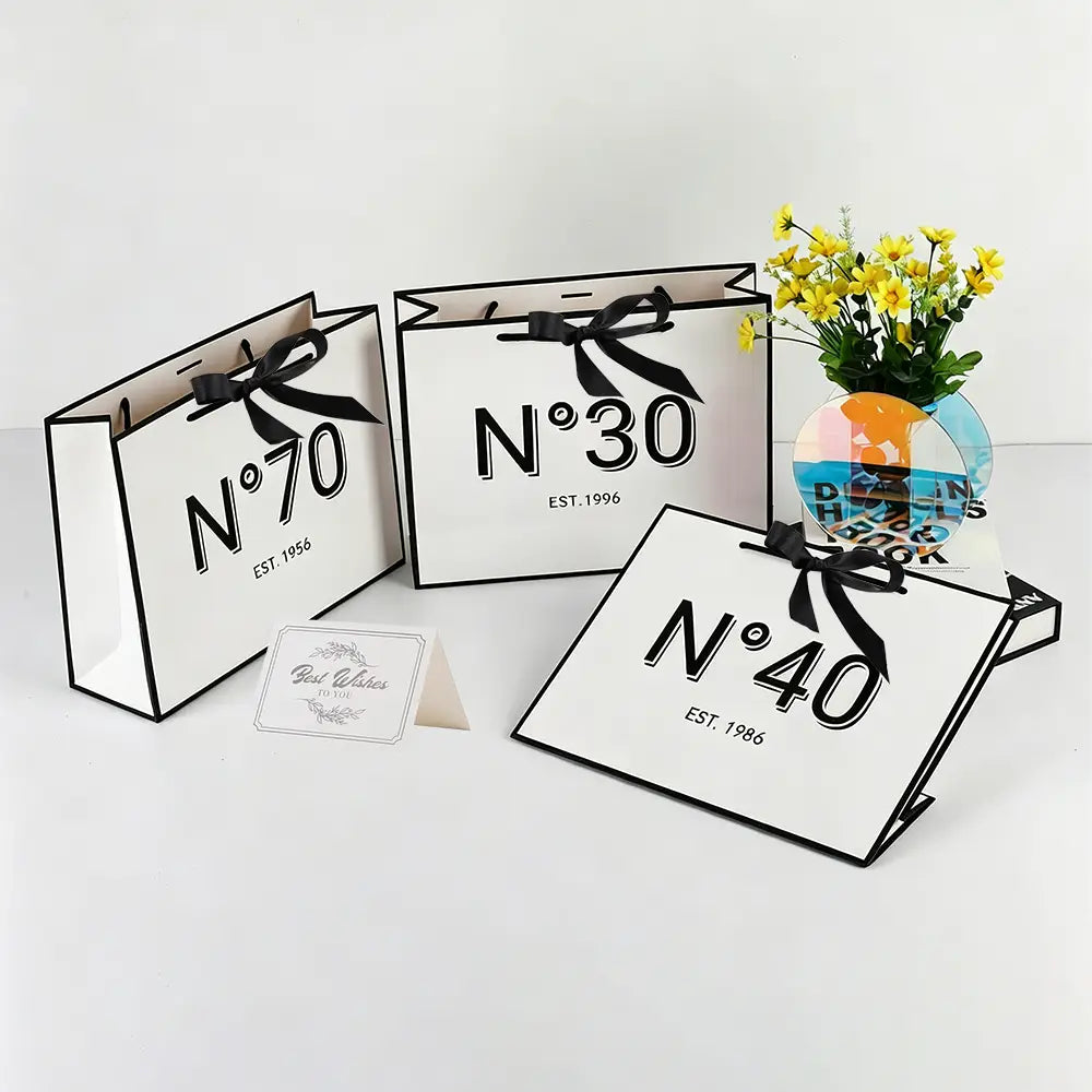 Tragetaschen aus weißem Papier mit schwarzen Schleifen und markanter schwarzer n-Nummerierung mit minimalistischer Gestaltung als Geschenkbeutel zum runden Geburtstag mit Karte und Schleife.
