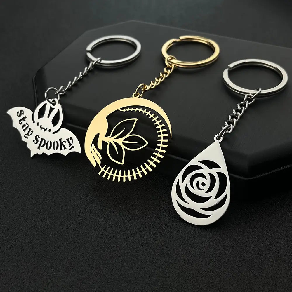 three custom engraved metal keychains featuring a silver bat charm engraved stay spooky a gold circular pendant with stitched leaf details and a white teardrop-shaped rose cutout designed as personalized gifts