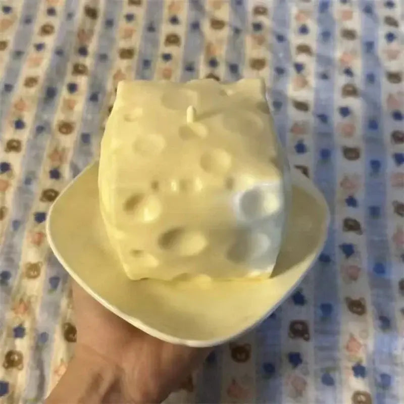 Un juguete antiestrés esponjoso con forma de queso, color blanco cremoso y marfil, con una mecha en la parte superior.