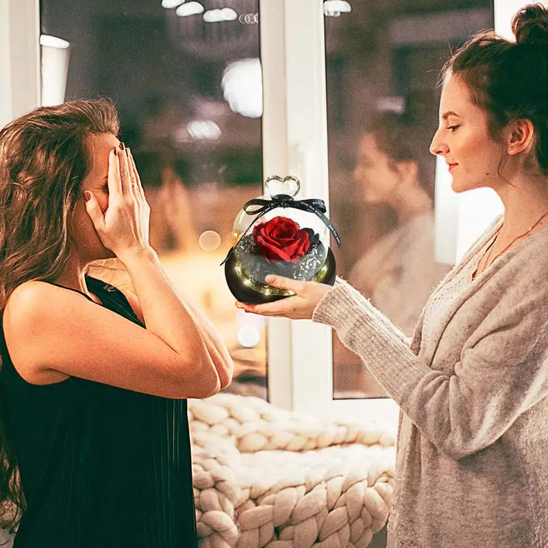 Une seule rose rouge préservée éclatante exposée à l’intérieur d’un dôme en verre transparent, avec un ruban noir attaché en forme de nœud cœur en haut, dans le cadeau en dôme en verre avec rose préservée et éclairage à LED.