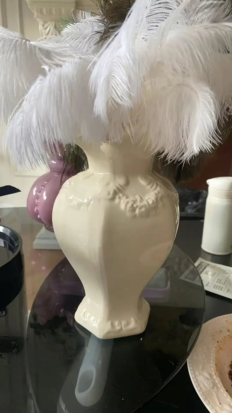 Jarrón de cerámica moderno francés color crema con ornamentación detallada en relieve y un cuello acampanado lleno de abundantes plumas blancas, para una decoración elegante del hogar.