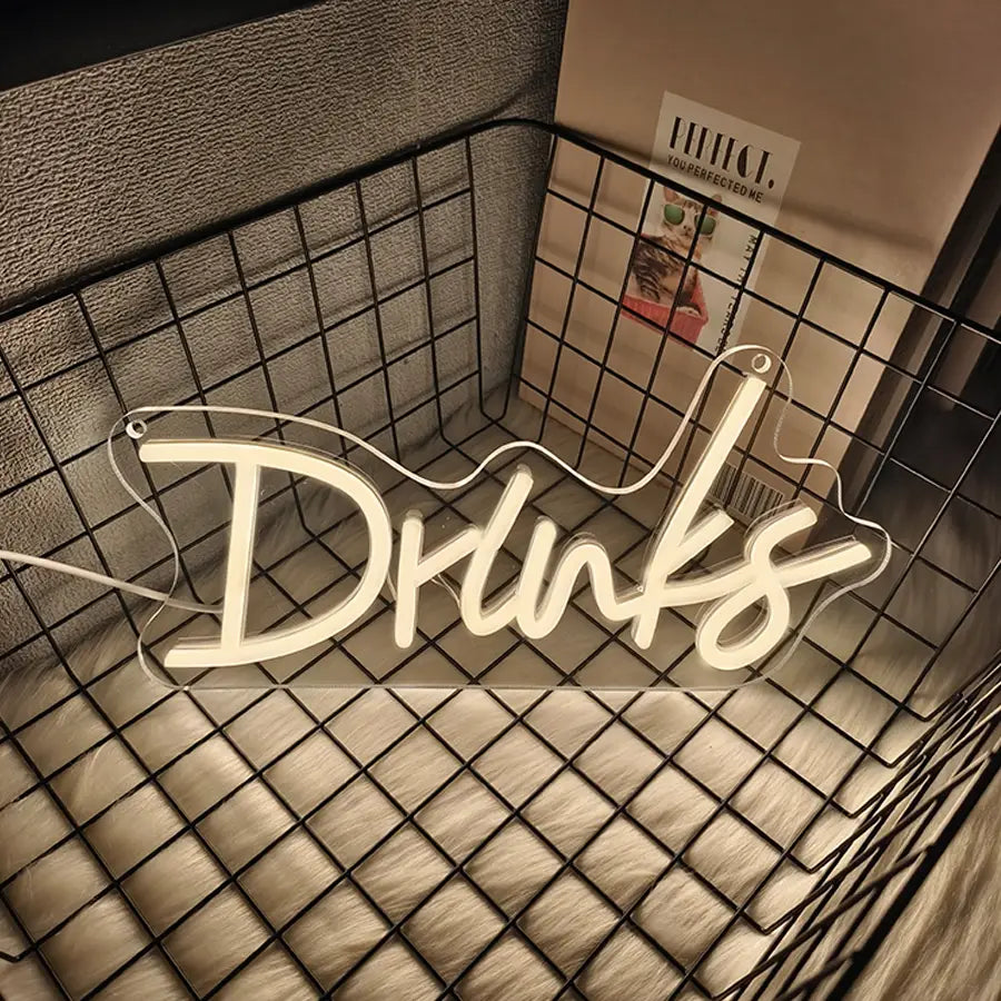 une enseigne au néon blanc lumineux épelant « drinks » dans une écriture cursive à LED, posée dans un panier en fil noir, comme décoration murale à LED pour enseigne au néon « drinks » en cursive