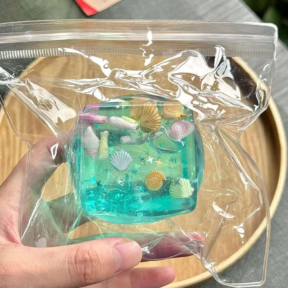 Une boîte de conservation en résine turquoise translucide remplie de mini coquillages, d’étoiles de mer et de charmes sur le thème de la mer, conçue comme un cadeau attentionné pour un cube sensoriel à effet anti-stress pour l’océan.