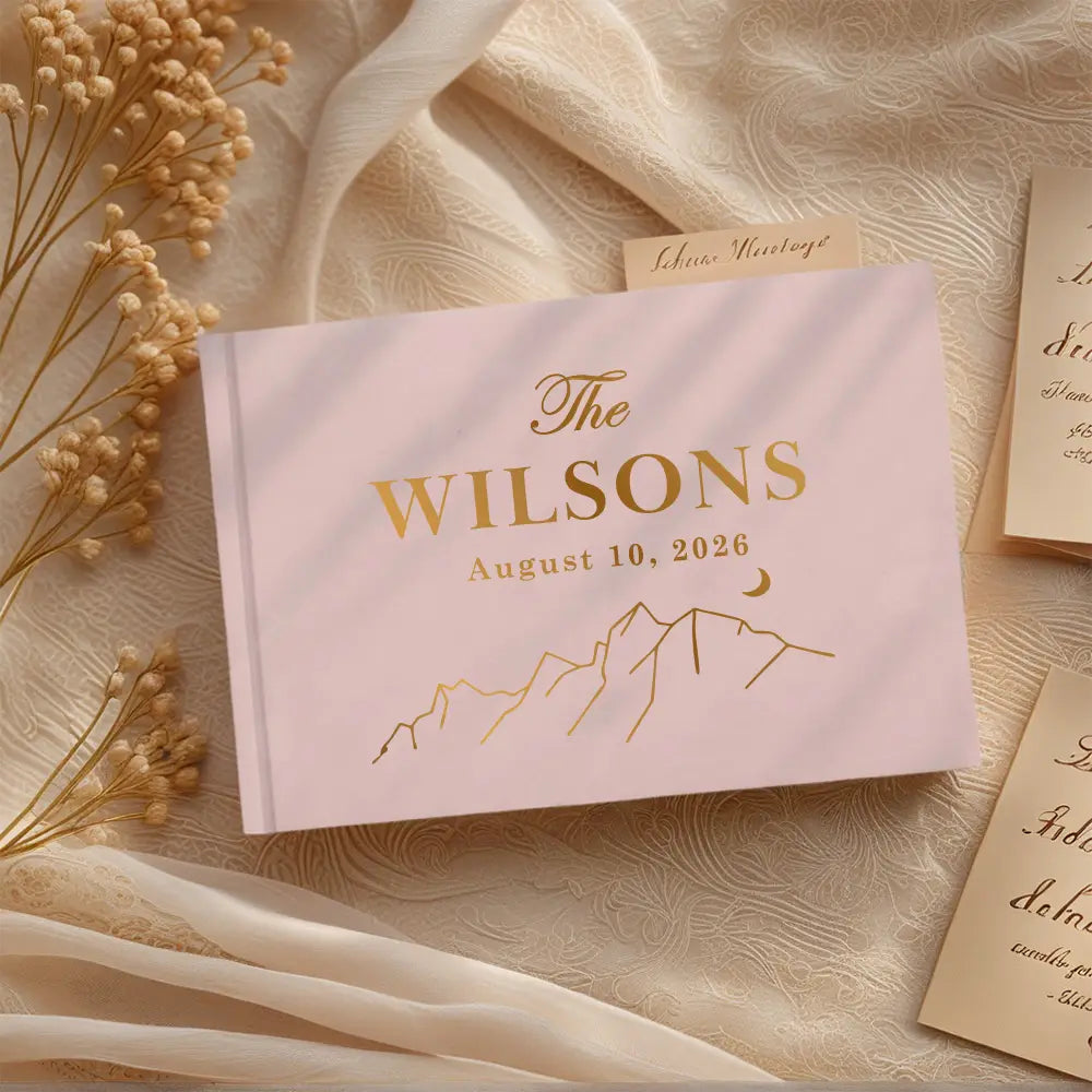 A soft pink personalized wedding guest book with gold foil lettering and a mountain design accompanied by a pen.