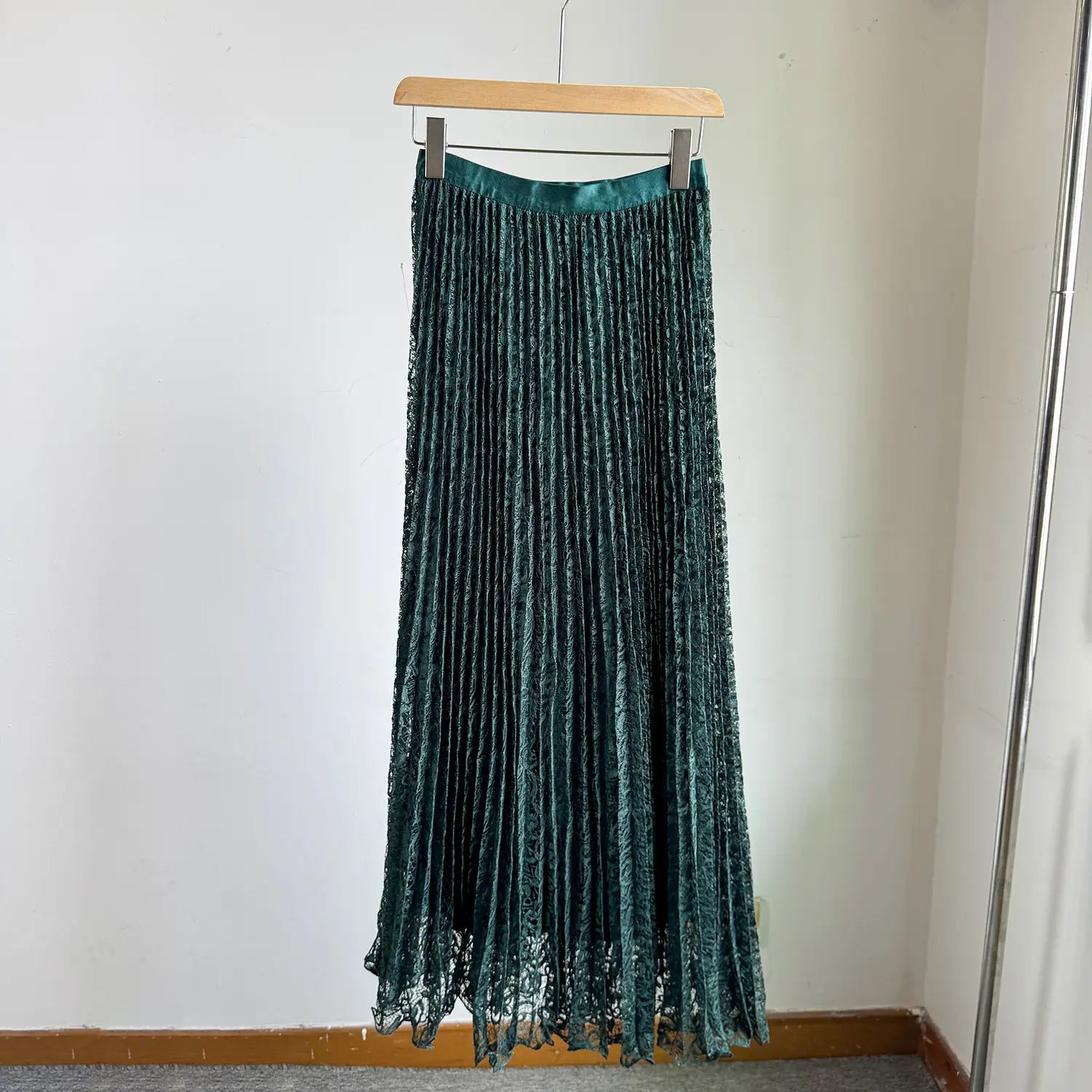 A high waist pleated midi skirt in deep emerald green velvet with a lace hemline hanging on a wooden hanger for Y2k streetwear.