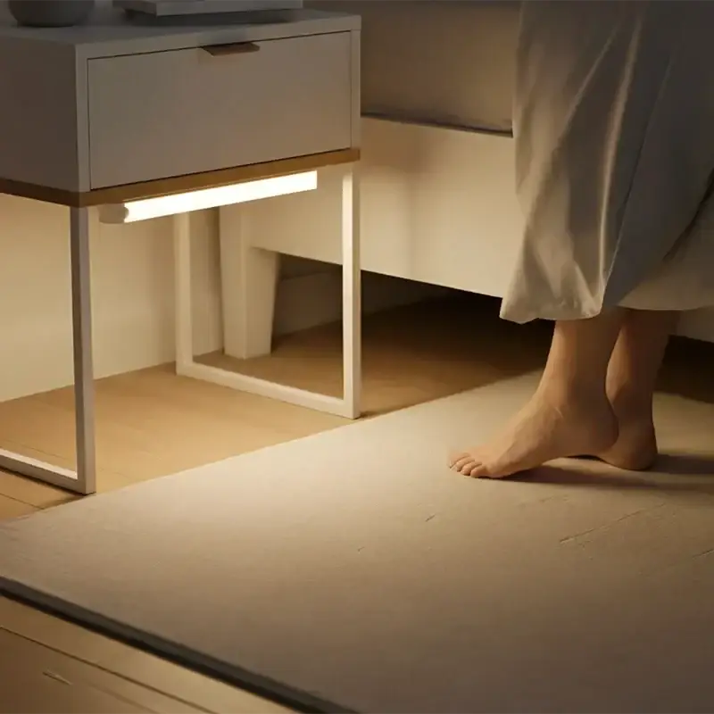 Una elegante mesita de noche blanca con diseño minimalista y un tirador de cajón con acentos dorados, que incorpora una lámpara lineal LED recargable por USB con sensor de movimiento a lo largo de su base, vista desde el frente