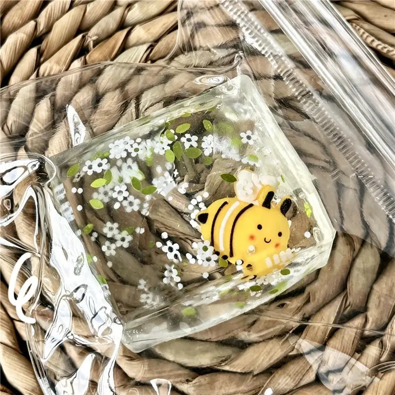 une figurine de petite abeille de dessin animé jaune et noire à l’intérieur d’un cube sphérique carré transparent en résine à faible rebond, rempli de fleurs blanches et de feuilles vertes, vue de face