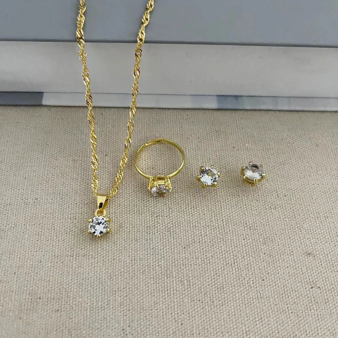 Juego de joyería de 5 piezas con circonitas cúbicas de color dorado, que presenta un collar de cadena tipo cuerda trenzada con un dije central de diamante, aretes tipo botón a juego y un anillo ajustable adornado con piedras de circonita cúbica transparentes.
