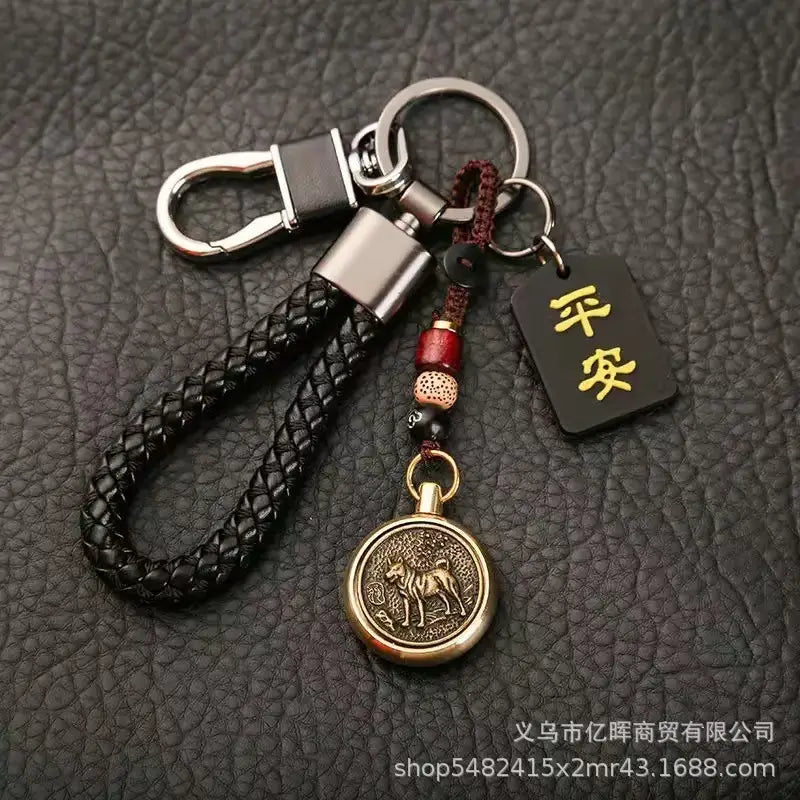 赤いビーズをアクセントにした黒の編みレザー製キーチェーン。ブロンズの「犬」メダリオン、シルバートーンの金属バックル、リングを備え、中国の干支をモチーフにした編みレザー製ラッキーペンダントのコレクションに含まれる、金色の漢字が刻まれた黒いタグ付き。