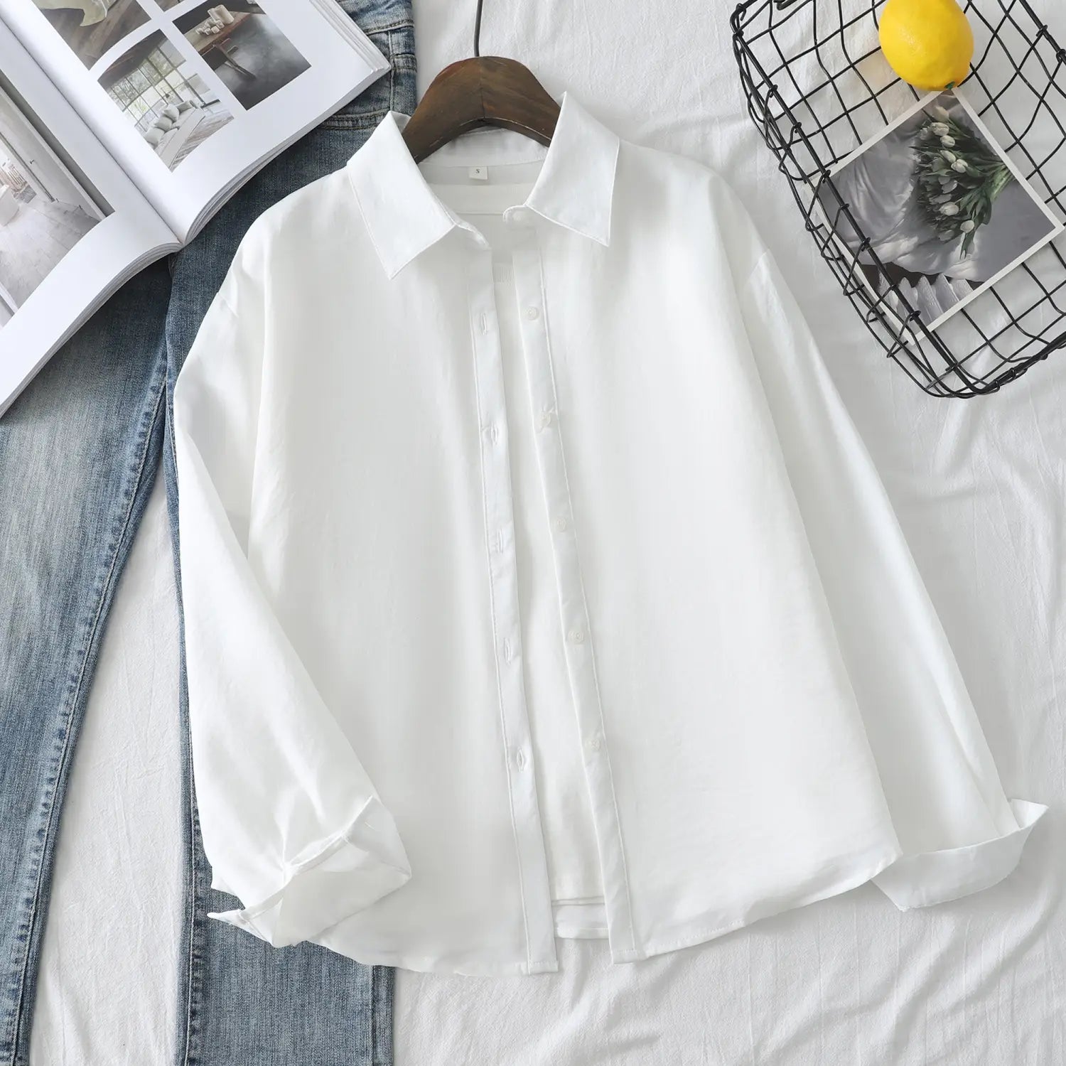 Eine knusprig-weiße Damen-Casualbluse aus Baumwollmischgewebe mit langen Ärmeln mit klassischem Kragen und dezenten Falten, ordentlich an einem Holzkleiderbügel aufgehängt.