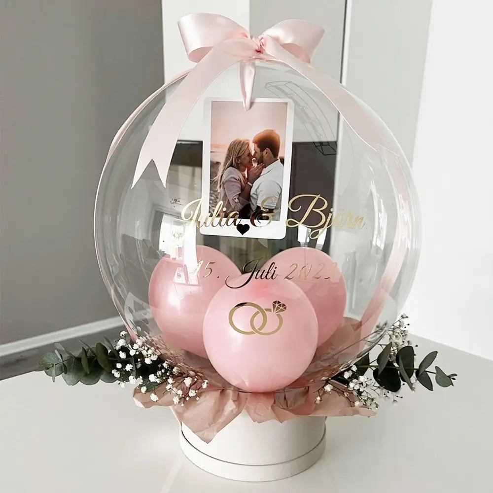 Eine klare, kugelförmige 36-Zoll-Bobo-Ballon-Domkonstruktion, geschmückt mit einer pinkfarbenen Satin-Schleife, die zarte rosafarbene Ballons und feines Grün umrahmt und ein Foto von zwei Personen zeigt, die sich küssen – individueller Baby-Party-Ballonaufkleber.