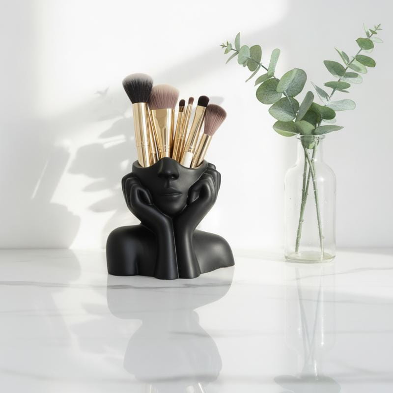 Black ceramic abstract face makeup brush holder with gold-tipped brushes nestled in its hollowed-out head functioning as a decorative vase.