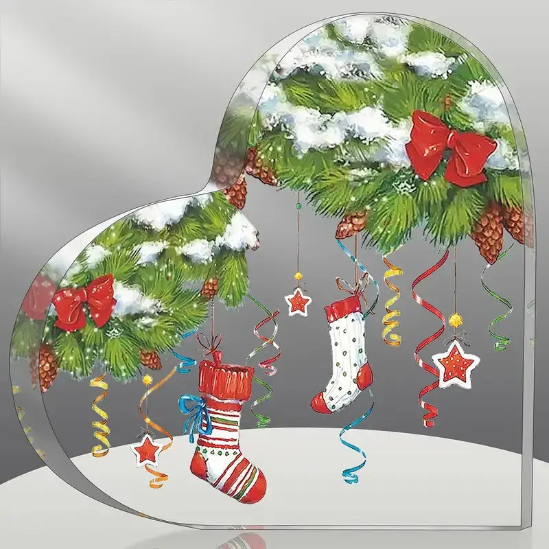Adorno de escritorio navideño de acrílico con forma de corazón, que presenta una escena festiva con ramas de pino verdes cubiertas de nieve, lazos rojos colgando, calcetines colgantes, cintas coloridas y decoraciones de estrellas.