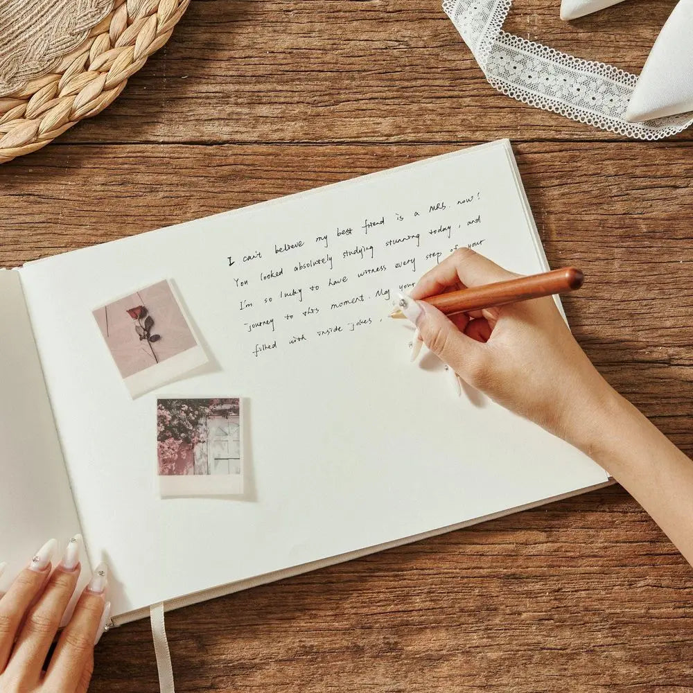 A hand holds a wooden pen writing on the white pages of an open cream-colored custom wedding guest book and pen linen photo album with two attached polaroid-style photos.