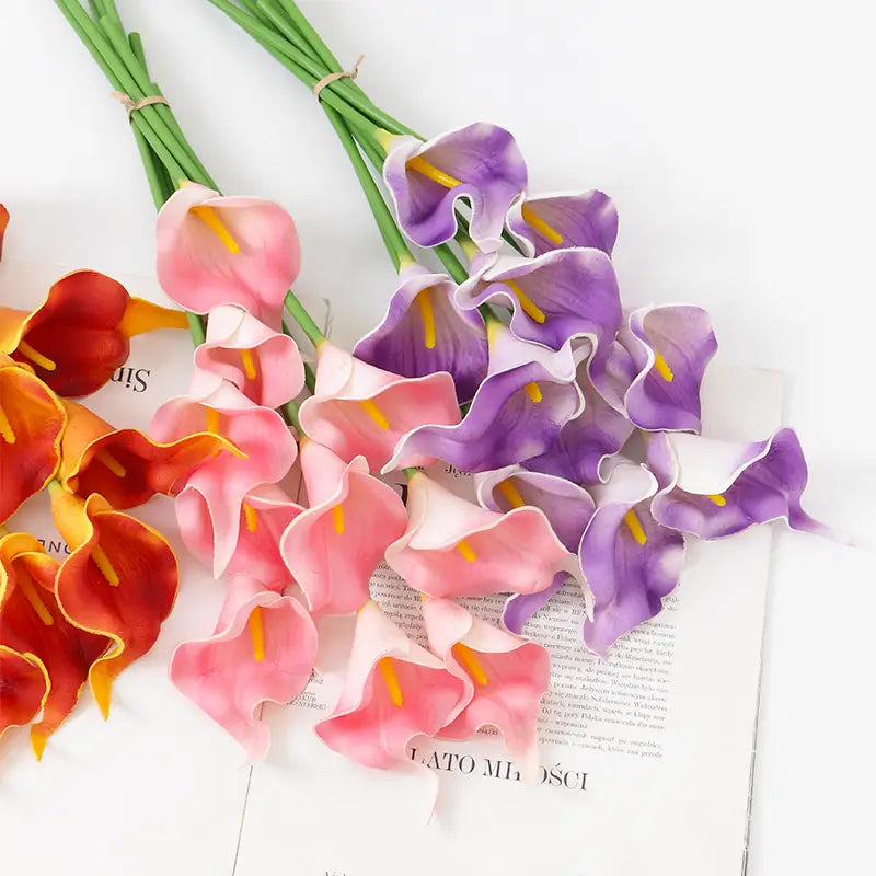 A bouquet of artificial calla lily flowers for home decor in soft pink, vibrant purple, and warm orange hues with yellow centers viewed from the front.