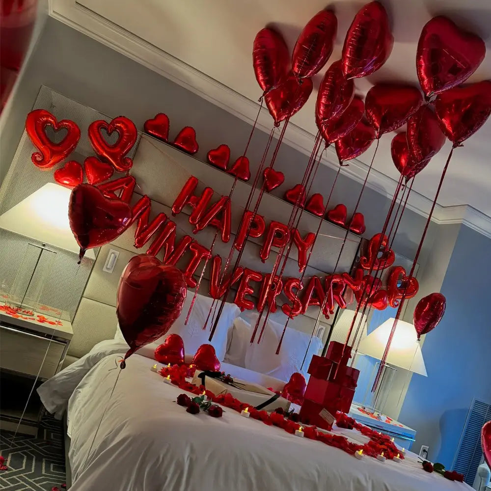 red heart-shaped foil balloons spelling happy anniversary arranged above a bed as part of an anniversary decoration set with rose petals