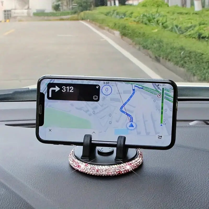 Un soporte negro para teléfono de automóvil con ventilación, de pedrería de color negro, con una base engalanada con pedrería brillante en rosa y transparente; sostiene un iPhone que muestra una aplicación de navegación.
