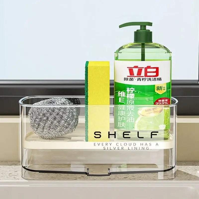 Organizador transparente de fregadero de cocina de plástico de dos niveles con sistema de drenaje que sujeta un frasco dispensador verde de jabón para platos, una esponja amarilla y un estropajo de alambre plateado.