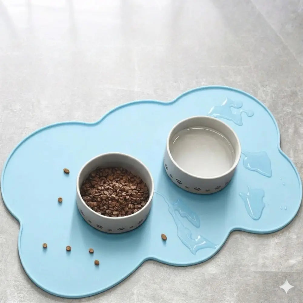 Alfombra de alimentación para mascotas de silicona con forma de nube color azul claro para perros y gatos pequeños y medianos, con dos cuencos de cerámica blancos con patrones de huellas; uno está lleno de alimento seco y el otro, de agua.