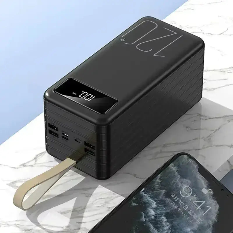 Black rectangular portable power bank with digital display showing 100 percent charge, multiple usb ports on the side, and a beige carrying strap attached.