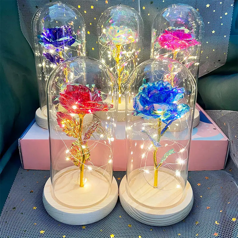 Bunte, glitzernde Rosen mit goldenen Stielen und eingebetteten Lichterketten, ausgestellt in klaren Glaskuppeln auf Holzsockeln, im „Eternal Gold Rose Glass Dome Led Light Gift“.