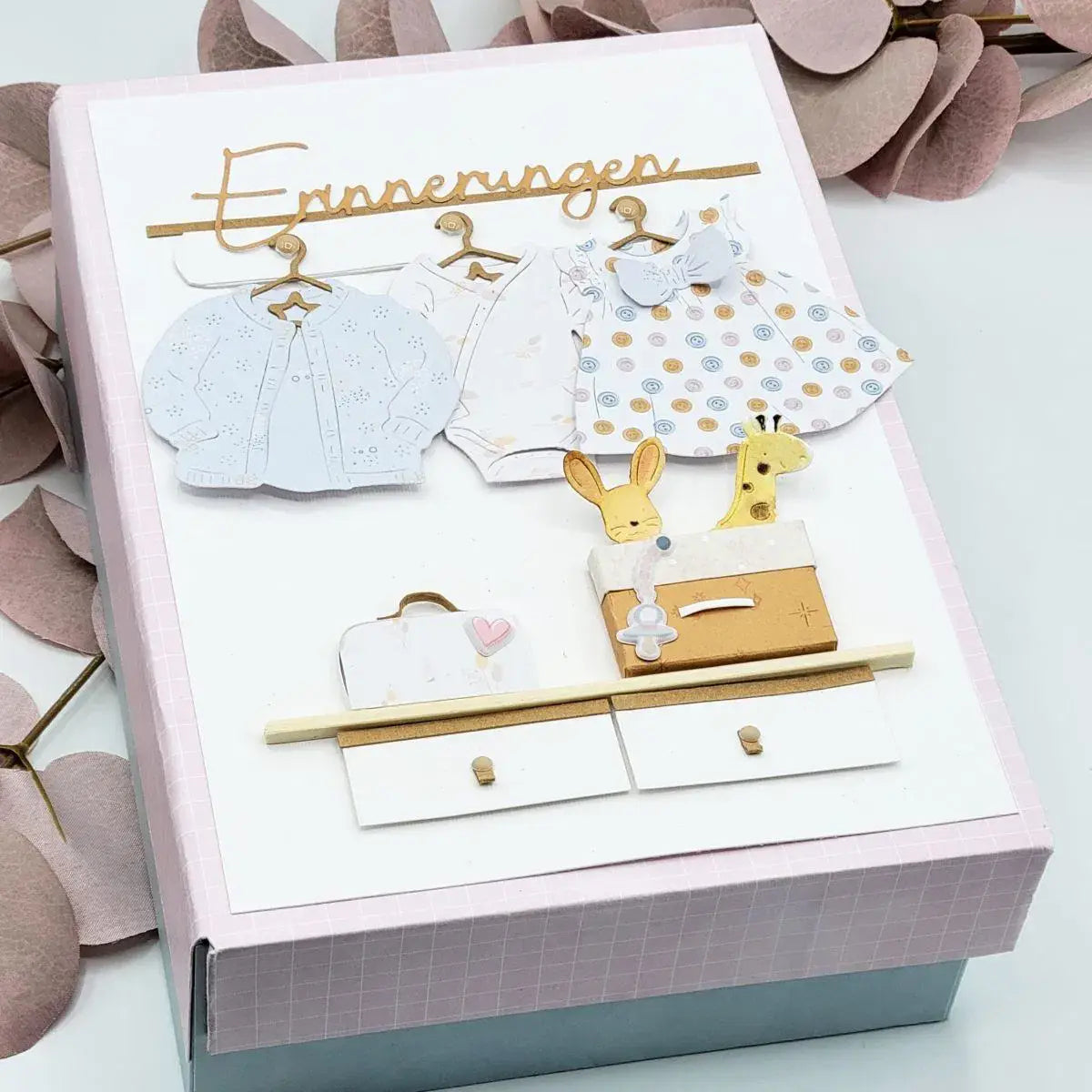 Eine zarte Karte-Box in Rosa und Weiß im Baby-Thema mit gestanzten Papierkleidungsstücken, goldenen Aufhängern und kleinen Tierfiguren, die auf einer Kommode präsentiert werden; hergestellt mit Oster-Ei-Hasen-Babyschneidvorlagen.