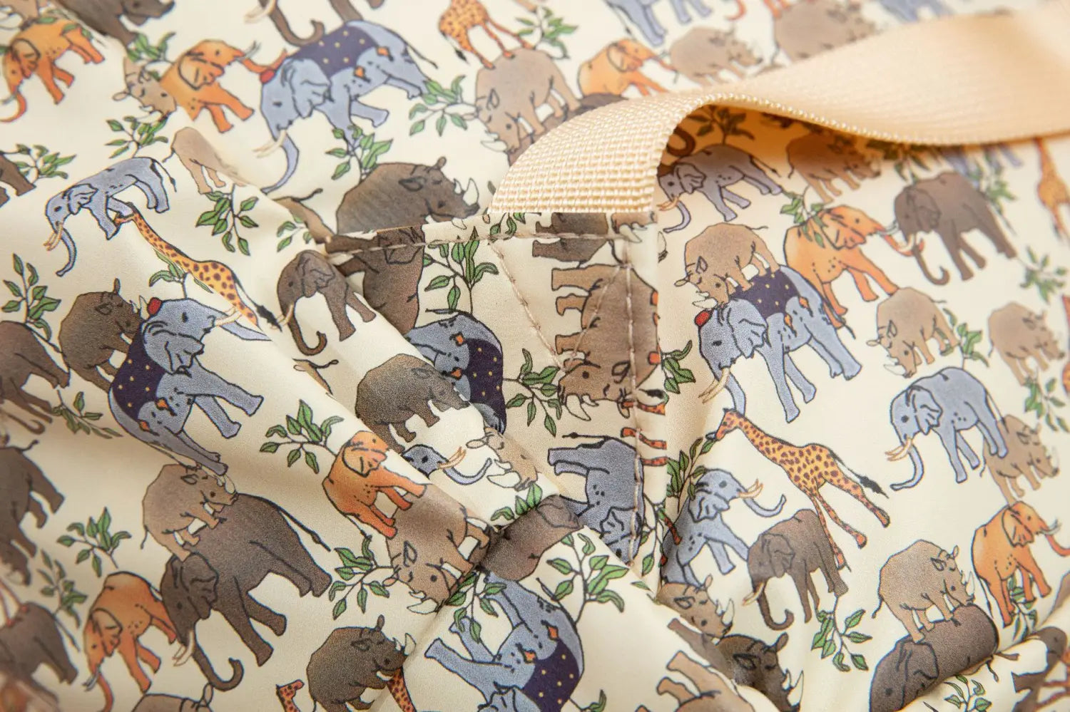 Sac à dos dinosaure pour enfants couleur crème, style sac d’école pastel, avec des éléphants, des girafes et des rhinocéros illustrés de manière ludique, aux nuances de bleu-gris, orange et brun, sur un support en tissu.