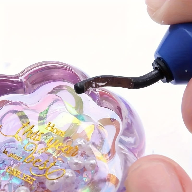 a translucent purple glass ornament with gold foil lettering being cleaned by a blue-handled resin epoxy trimming tool manual jewelry cutter.