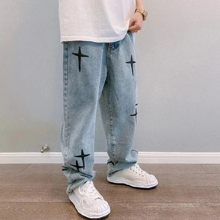 Jeans en denim délavé clair avec broderie croisée noire sur les jambes, présentant un design ample à jambes larges.
